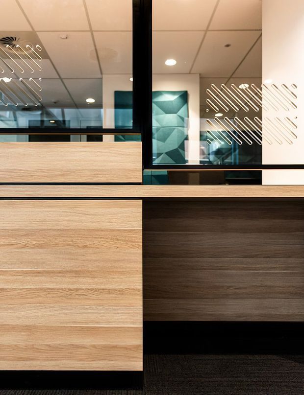 Close-up view of a contemporary built-in reception or storage joinery installation, featuring horizontal light timber cabinetry paired with darker wood panels and black trim. Large mirrored glazing above the joinery incorporates etched linear decorative markings and reflects a modern interior with geometric artwork. A suspended acoustic-tile ceiling and recessed lighting are visible in the reflection.