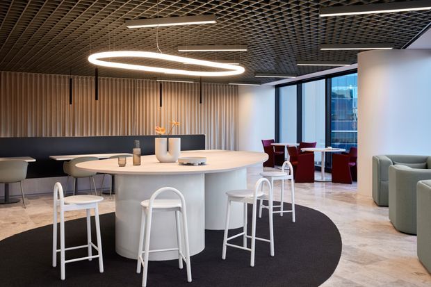 Contemporary commercial hospitality or breakout lounge with a large rounded white communal island table and white bar stools, surrounded by soft seating and small dining tables. The space features a dark acoustic grid ceiling, sculptural linear and circular pendant lighting, vertical timber wall panelling, dark upholstered banquette seating, polished light flooring, and floor-to-ceiling glazing overlooking an urban setting.