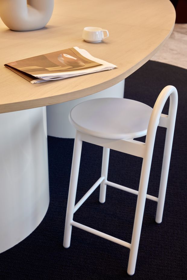 Close-up interior view of a softly rounded light-wood communal table with a white curved pedestal base, paired with a white high stool. A white ceramic vessel, cup, and magazine rest on the tabletop. The space has dark charcoal carpet and a clean, restrained contemporary aesthetic.