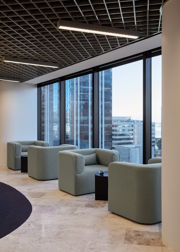A refined commercial lounge or waiting area with several rounded, upholstered armchairs arranged beside small dark side tables. The space features full-height city-facing glazing, pale stone flooring, white walls, and a distinctive exposed dark grid ceiling with linear suspended lighting. High-rise buildings are visible through the windows, reinforcing an urban office or hospitality setting.