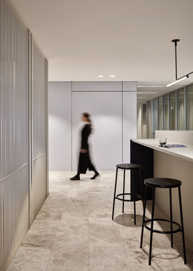 A refined contemporary commercial interior, likely an office reception or hospitality-style waiting area. The long space features pale stone-look tiled flooring, smooth white panelled walls, ribbed translucent screening along the left, extensive framed glazing on the right, and a minimalist counter with two black bar stools. Linear and recessed lighting create a clean, softly illuminated atmosphere.