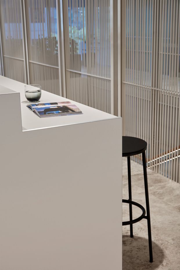 A minimalist interior vignette showing a crisp white built-in island or counter with a slim white benchtop, a black bar stool, a glass, and a magazine. Behind it is a full-height vertical slatted screen or partition with glazed openings and daylight beyond. The floor appears to be pale polished concrete or large-format stone tile. The restrained palette and clean geometric lines create a refined contemporary atmosphere.