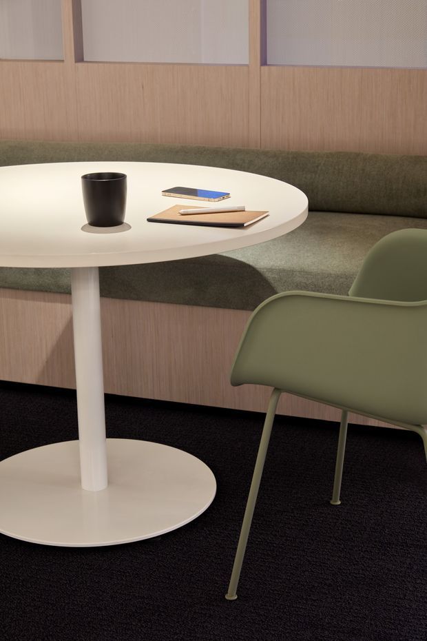 A softly lit contemporary interior seating area with a round white pedestal table, olive-green upholstered chair, and built-in curved banquette. The space features warm vertical timber wall panelling, green upholstery, white acoustic or framed wall panels, and dark carpet. A black cup and notebooks rest on the table, suggesting a refined office, lounge, or collaborative workspace.