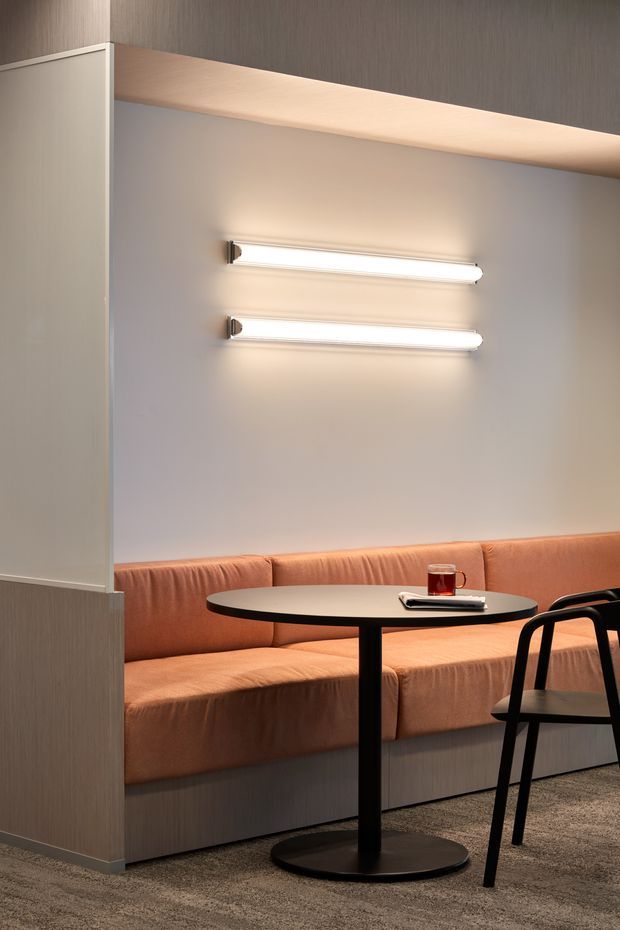 A softly lit contemporary hospitality-style booth seating area with a built-in peach-beige upholstered banquette, round black pedestal table, and black chairs. The booth is partially enclosed by a pale wood-toned partition and overhead canopy, with two horizontal wall-mounted tube lights creating a warm glow. Neutral carpet and restrained detailing give the space a refined, intimate feel.