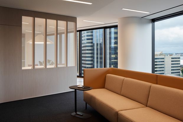 Contemporary office lounge or reception area with a large tan upholstered sectional sofa, slim black side table, dark carpet, and a timber-toned glazed partition. Floor-to-ceiling windows provide an urban city view, while a cylindrical white column and linear recessed lighting reinforce the clean commercial interior.