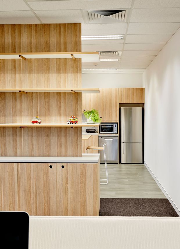 Bright contemporary kitchenette and breakout area in a commercial office, featuring extensive light timber veneer cabinetry and open shelving, integrated microwave and dishwasher, a stainless-steel refrigerator, pale grey flooring, and a white suspended acoustic-tile ceiling with fluorescent lighting. A small breakfast counter with stools and a partially visible upholstered seating area occupy the foreground.
