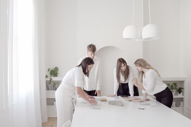 Bright, minimalist white-toned interior showing a group of people collaborating around a large white table with architectural or design plans and materials. The room features floor-to-ceiling sheer curtains, pale walls, simple white globe pendant lights, built-in shelving, and subtle indoor greenery.