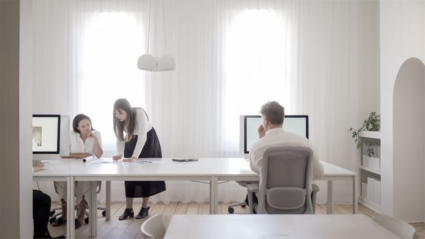 Bright, minimalist office or studio interior with a long white shared worktable, three people collaborating, office chairs, paperwork, and soft pendant lighting. Tall sheer-curtained windows provide abundant daylight, while an arched wall opening and simple white shelving with a plant add subtle architectural detail. Pale timber flooring and predominantly white finishes create an airy, calm workspace.