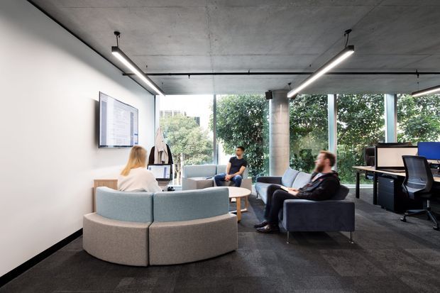 A contemporary open-plan office lounge and workspace with curved upholstered seating, armchairs, a small round coffee table, workstations, and large floor-to-ceiling windows overlooking greenery. The space features a raw exposed concrete ceiling and column, dark carpet flooring, white walls, and linear suspended lighting.