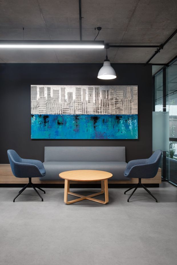 A modern reception or lounge area with a grey sofa, two blue upholstered armchairs, and a round light-wood coffee table. The space features polished concrete flooring, a dark painted feature wall, exposed concrete ceiling, black-framed glazing, suspended linear and pendant lighting, and a large abstract artwork in blue and white.
