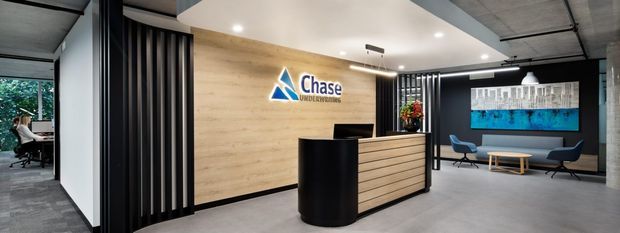 Modern commercial reception area with a black curved reception counter and timber-fronted desk, a light timber feature wall displaying the Chase Constructions logo, black vertical screening panels, polished concrete flooring, exposed concrete ceiling, and a small seating area with artwork in the background.