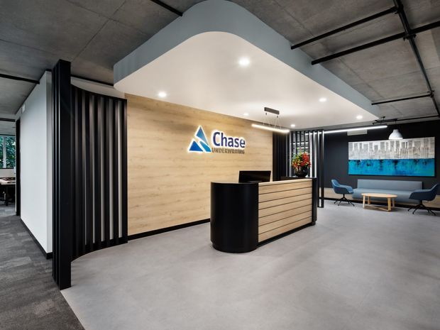 A polished commercial office reception area with a curved white bulkhead, exposed concrete ceiling, black vertical screening, pale timber feature wall, and a two-tone reception desk bearing the Chase Underwriting logo. A small waiting area with blue chairs, a timber coffee table, artwork, and greenery is visible in the background.
