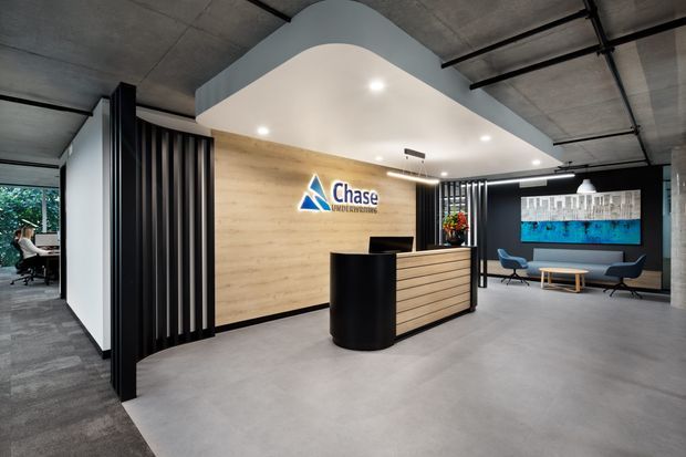 Contemporary commercial reception area with a curved white ceiling bulkhead, exposed concrete ceiling, polished concrete floor, timber feature wall, black vertical slatted screens, and a black-and-timber reception desk. A corporate logo is mounted on the feature wall, with a small waiting area and artwork visible in the background.