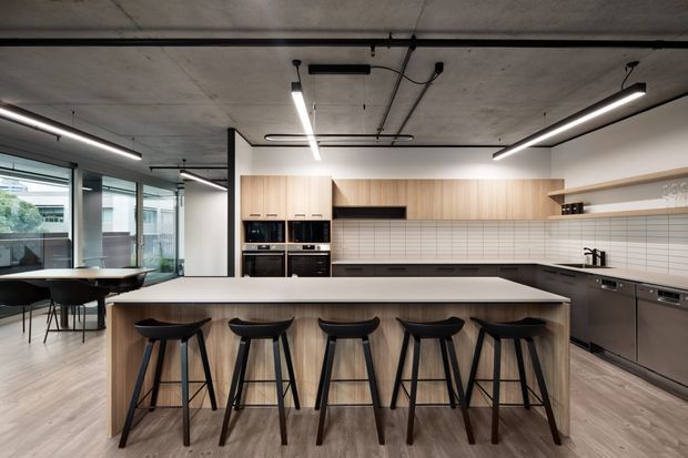 A spacious contemporary kitchen with a long light-wood island and five black bar stools, dark cabinetry, integrated appliances, and a white tiled backsplash. The room features exposed concrete ceilings, black linear pendant lighting and track services, large glazed windows, and an adjacent dining area.
