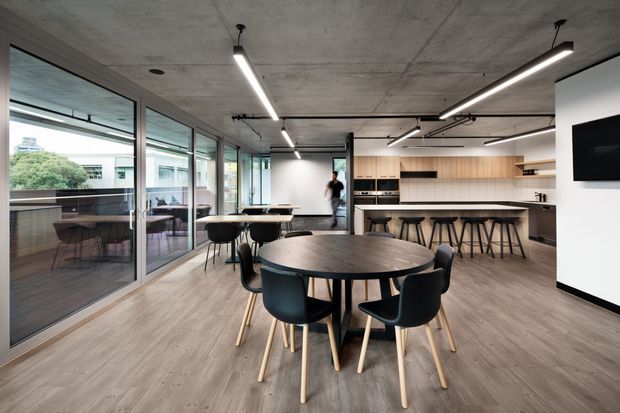 Contemporary open-plan office or hospitality-style breakout space with a large communal dining table in the foreground, a long island bench with bar stools and light timber cabinetry at the rear, and extensive floor-to-ceiling glazing opening onto an outdoor balcony. The room features an exposed concrete ceiling, linear suspended lighting, grey timber-look flooring, black-framed glazing, and a wall-mounted television.