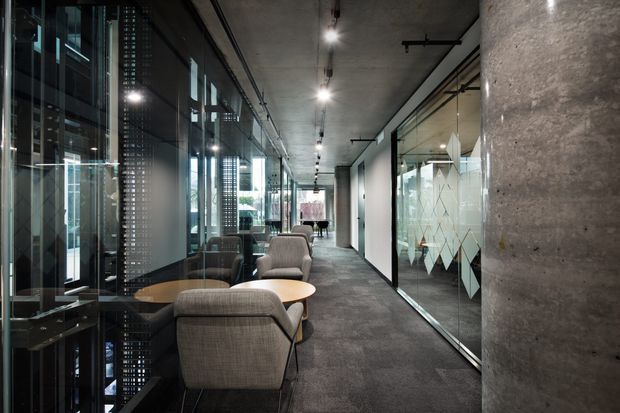 A contemporary commercial office corridor with exposed concrete columns and ceiling, dark carpet flooring, extensive glazed partitions, and a row of lounge seating and small timber tables along the window side. Linear track lighting and visible services reinforce the industrial office character.