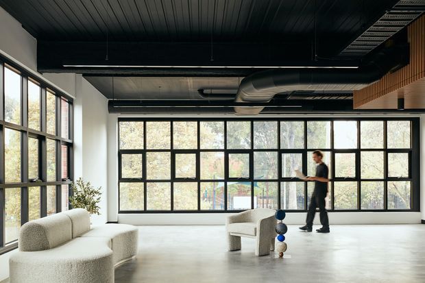 A spacious contemporary interior with expansive black-framed industrial-style glazing, a dark slatted ceiling, exposed black ventilation ducts, and a polished light floor. Minimal upholstered seating and a sculptural side table create an open lounge or commercial studio atmosphere, with a person walking near the windows.