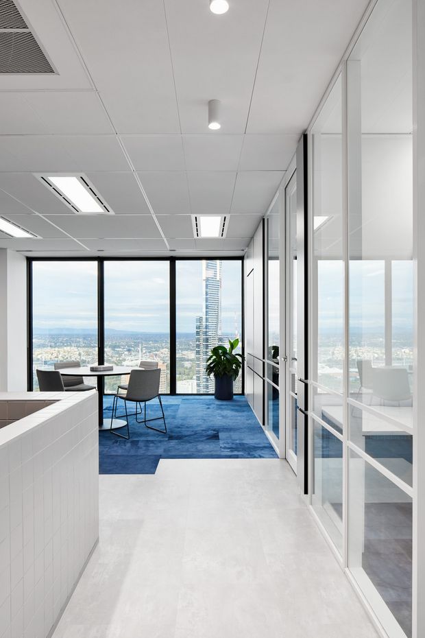 A bright contemporary commercial office interior with a white tiled reception or kitchenette counter in the foreground, white acoustic panel ceiling with recessed linear lights, extensive glass-partitioned rooms along the right, and a small meeting area beside floor-to-ceiling windows. Blue carpet defines the meeting zone, with a potted plant and expansive city views beyond.