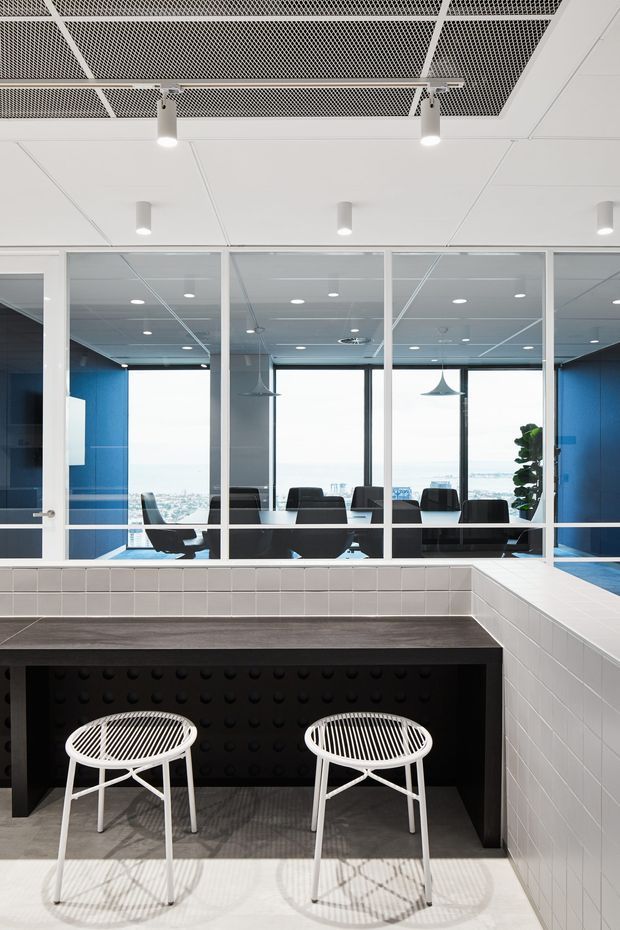 Contemporary commercial office interior featuring a black and white tiled reception or breakout counter in the foreground with two white stools. Behind it, a fully glazed meeting room with dark conference chairs is framed by vivid blue walls. The space has a white acoustic panel ceiling, exposed black mesh ceiling panels, track-style spotlights, pendant lights, and extensive city-facing glazing.
