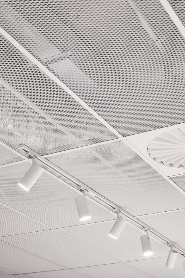 Close-up upward view of a bright white commercial-style ceiling with perforated acoustic ceiling panels in a regular grid. Several white track-mounted cylindrical spotlights run diagonally across the frame, and a circular ceiling ventilation or diffuser element is visible to the right. The image is minimal and monochromatic, focused on ceiling construction and lighting.