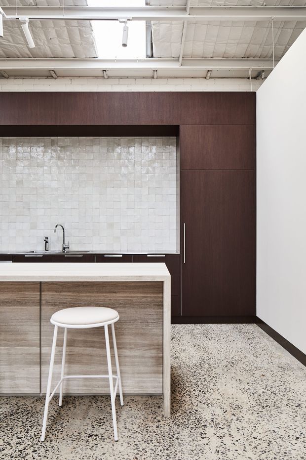 A refined contemporary kitchen with dark timber cabinetry, a pale textured square-tile splashback, a light stone island benchtop, and a white stool. The kitchen sits within an industrial-style shell featuring an exposed white-painted ceiling, visible services, and a polished terrazzo floor.