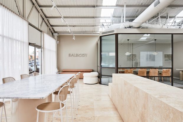 Bright contemporary commercial hospitality or workplace interior with a long pale stone communal table, curved timber stools, a large stone service counter, glass-partitioned meeting room, and exposed industrial ceiling services. The space has abundant daylight through sheer vertical blinds and a refined neutral palette.