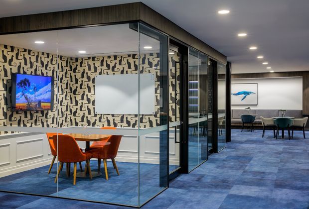 A contemporary commercial interior with a glazed meeting room enclosed by black-framed glass partitions. Inside, a small timber meeting table is surrounded by orange upholstered chairs, with white wall panelling, patterned feature wallpaper, a wall-mounted screen and a whiteboard. The surrounding office has blue carpet, dark timber ceiling bulkheads, recessed lighting, and a furnished lounge or breakout area visible beyond.