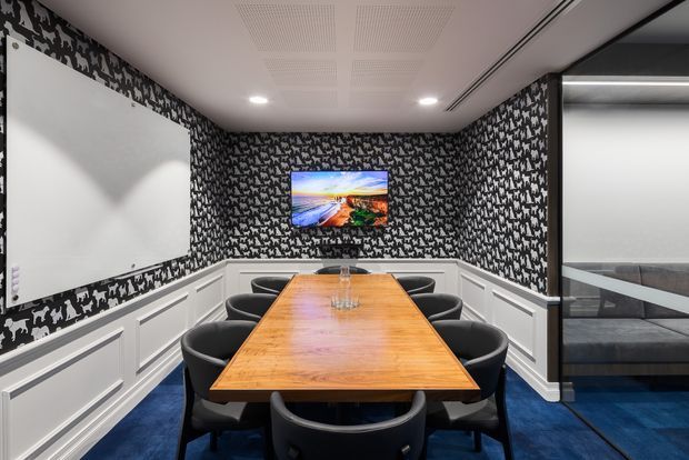 A compact, enclosed meeting or boardroom with a long warm timber conference table surrounded by black chairs. The room features striking black-and-white patterned wallpaper, white decorative wall panelling, a wall-mounted screen, acoustic ceiling panels, recessed lighting, and deep blue carpet. Glazing on the right reveals an adjacent upholstered seating area.