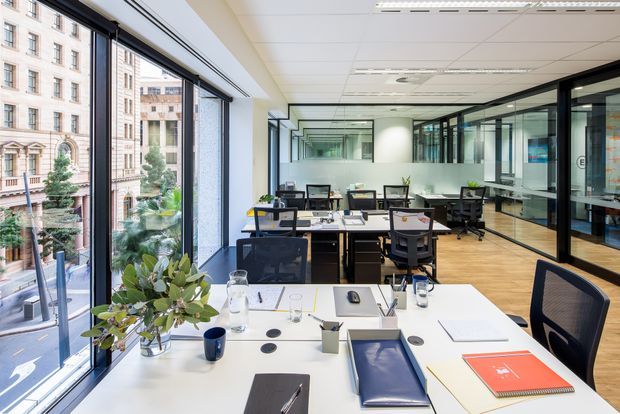 Bright contemporary open-plan office with white workstations, ergonomic task chairs, computers, office supplies, and indoor plants. Full-height glazing along one side overlooks an urban streetscape, while black-framed glass partitions create enclosed meeting or work areas. The space has a suspended acoustic ceiling, light timber flooring, and a clean professional fit-out.