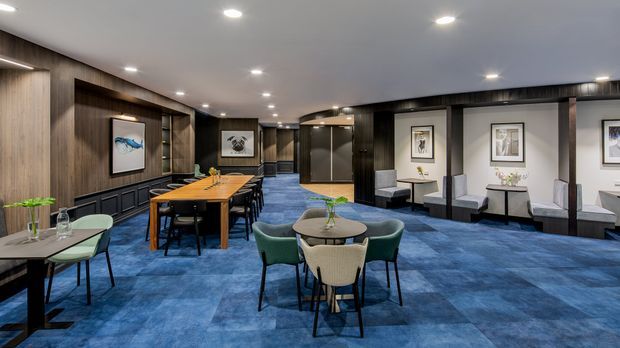 A spacious, contemporary commercial lounge or hospitality interior with deep blue carpet, dark timber wall panelling, recessed downlighting, and a series of seating and dining areas. A long timber communal table with black chairs occupies the left side, while small round tables and upholstered chairs are arranged throughout. Framed artwork, console tables, and a curved glazed partition add visual interest. The space has a refined, modern hotel or office-lounge character.