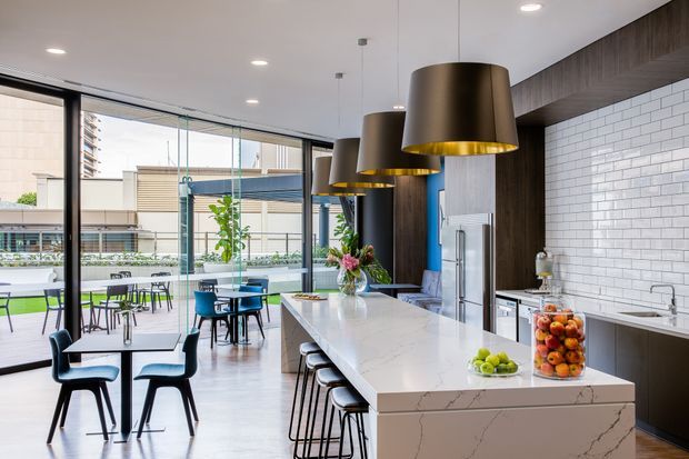 A bright contemporary kitchen and dining space with a large white stone island, integrated seating, dark timber cabinetry, white subway-tile splashback, stainless-steel appliances, and three oversized bronze pendant lights. Full-height glazing opens onto an outdoor courtyard or terrace with additional dining furniture.