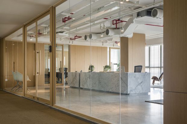 A contemporary commercial reception area viewed through a full-height glazed partition. The space features a large marble reception desk, light timber wall panelling and door frames, polished concrete flooring, exposed services and ductwork in the ceiling, and black-framed office furniture. The palette is light and refined with an industrial ceiling treatment.