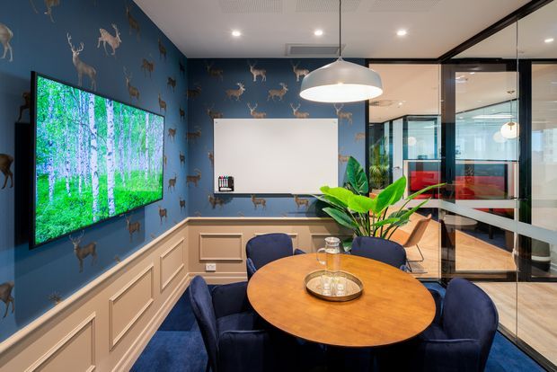 A stylish meeting or dining room with a round timber table surrounded by dark blue upholstered chairs. The room features navy deer-patterned wallpaper, blue carpet, white built-in wall panelling and cabinetry, a large wall-mounted display, a projector screen, indoor plants, and black-framed glass partitions opening to a bright adjacent office area. A large white pendant light hangs over the table.
