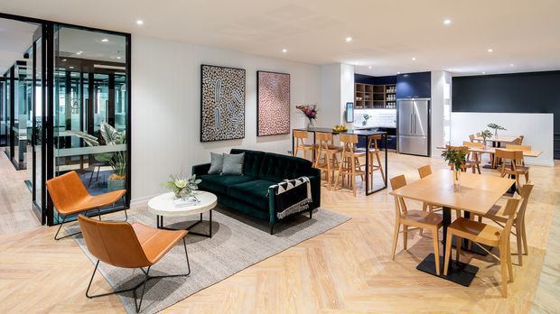 Open-plan contemporary living, dining and kitchen area with herringbone timber flooring, a dark green sofa, tan leather lounge chairs, timber dining tables and chairs, and a compact kitchen with dark cabinetry and stainless-steel appliances. Black-framed glazed doors create a visually connected enclosed space at left, while abstract wall art and plants add decorative detail.