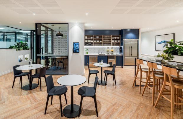 A bright contemporary commercial hospitality or office breakout space with a café-style kitchenette and bar at the rear, dark blue cabinetry, open shelving, white tiled splashback, stainless-steel refrigerator, and multiple small round tables with mixed seating. The room features warm herringbone timber flooring, a white ceiling with acoustic panels and recessed lighting, glazed meeting-room partitions, indoor plants, and a long timber communal counter with stools.
