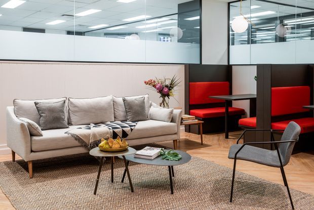 A contemporary office breakout and lounge area with a large upholstered sofa, patterned rug, nesting coffee tables, and an armchair in the foreground. Behind the lounge is a casual meeting or dining zone with built-in red banquette seating, dark partitions, tables, and glass-fronted office spaces. The space has a suspended grid ceiling, pale timber flooring, neutral wall finishes, and pendant globe lights.