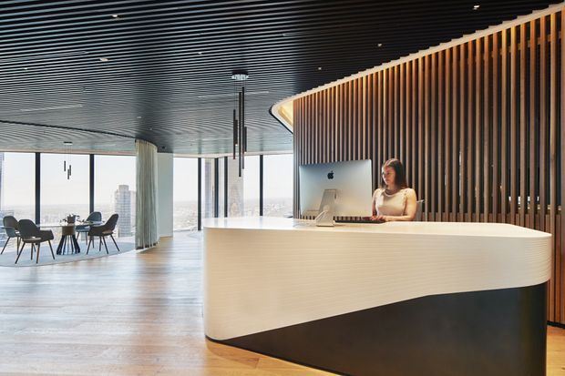 A spacious contemporary commercial reception area with a sculptural curved reception desk in the foreground. The desk has a white textured face and dark lower panel, backed by a vertical timber-slat feature wall. A receptionist works at a computer behind the counter. The space includes a dark linear slatted ceiling, polished timber flooring, floor-to-ceiling glazing, a small seating area, and expansive city views.
