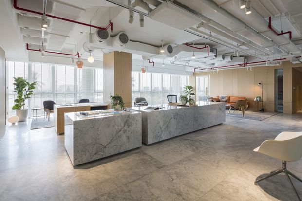 Bright open-plan contemporary commercial interior with two large marble-clad island counters, a dining or collaboration table, lounge seating, extensive floor-to-ceiling glazing, and exposed building services. The ceiling has exposed white-painted structure and ducts with visible red fire-sprinkler piping and track lighting. The space is furnished in a restrained neutral palette with timber wall panels, concrete-look flooring, greenery, and panoramic urban views.