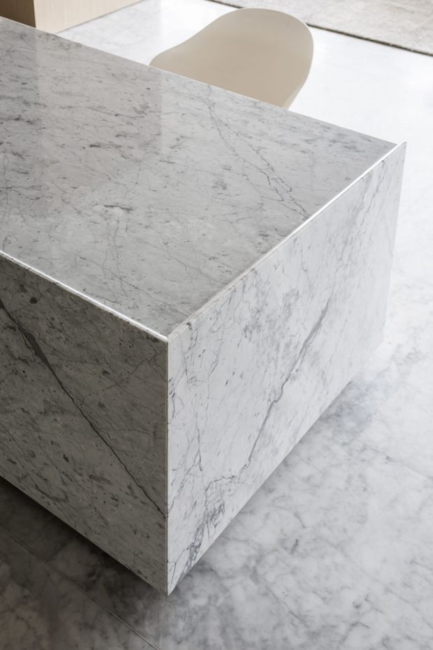 Close-up of a minimalist stone-clad kitchen island or counter, with continuous pale grey veined marble across the benchtop and waterfall sides. A light beige curved dining or bar chair is partially visible behind it, and the surrounding floor appears to be matching marble or marble-look stone. The composition emphasizes clean geometric edges, refined natural stone veining, and a neutral contemporary interior.