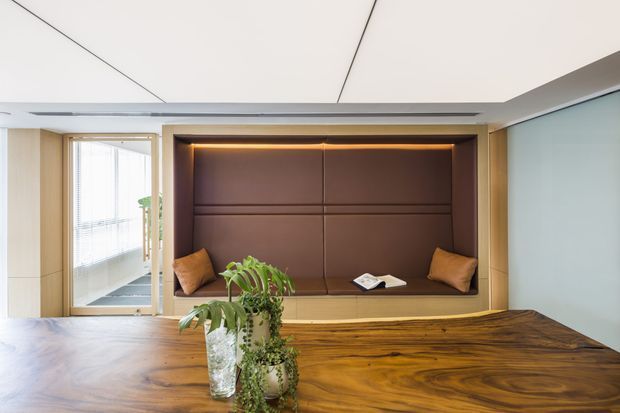 A refined contemporary interior featuring a built-in window seat or lounge nook with dark brown padded wall panels, warm concealed lighting, and timber cabinetry. A large richly grained timber table occupies the foreground, with potted greenery and small cushions adding warmth. Pale timber wall joinery, a glazed door, and a clean segmented white ceiling complete the space.