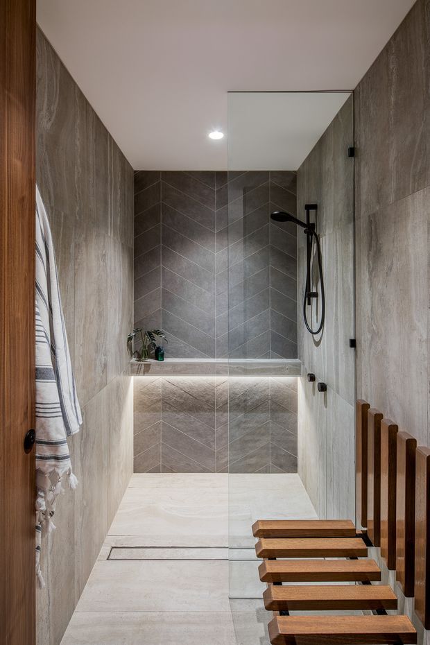 A compact contemporary walk-in shower room with full-height grey stone-look wall tiles arranged in a chevron pattern, pale stone-look floor tiles, a frameless glass shower screen, black shower hardware, and a floating recessed shelf with integrated lighting. A timber slatted bench and matching vertical timber detail line the entrance, with a striped towel hanging at the side.