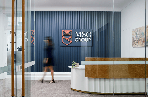 A contemporary commercial reception area viewed through glass partitions. The space features a curved reception desk with a white vertically ribbed front and warm timber-toned counter and fascia, a dark blue vertically fluted feature wall with the MSC Group logo, neutral carpet, and framed artwork. A blurred person is walking through the lobby.