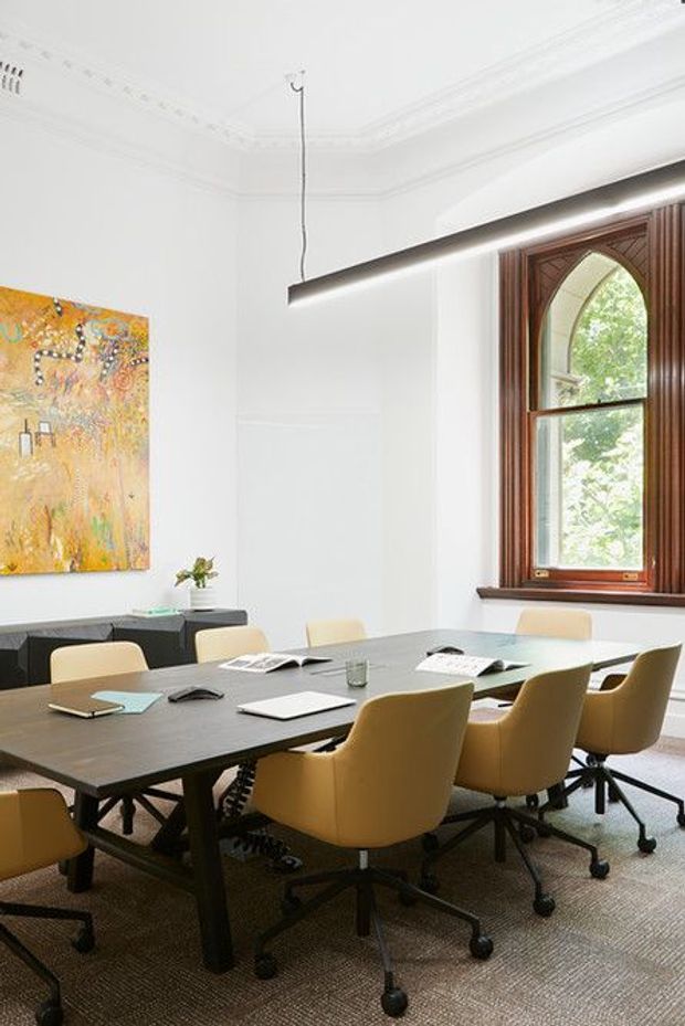 A refined meeting room or boardroom in a heritage-style interior, featuring a large dark timber conference table surrounded by tan upholstered swivel chairs. White walls with ornate cornices and ceiling detailing contrast with a richly stained timber window surround and pointed-arch window design. A slim suspended linear pendant light hangs above the table, while an abstract artwork and low dark sideboard add visual interest. The room has a professional, warm, contemporary fit-out within a traditional architectural shell.