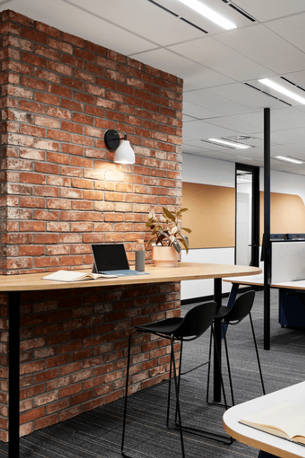 A contemporary open-plan office interior with a long light timber work counter mounted against an exposed red brick wall. Black bar stools, a laptop, books and a potted plant sit along the counter. The space includes black-framed glazed partitions, dark carpet, black structural columns, a white suspended acoustic ceiling with linear lighting, and a warm tan feature wall in the background.
