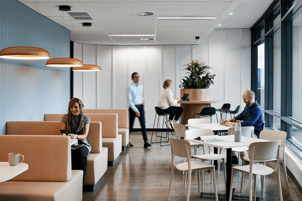 A bright contemporary commercial breakout and dining area with upholstered booth seating, café tables and chairs, a high communal table, pendant lighting, indoor planting, polished concrete flooring, and full-height windows.