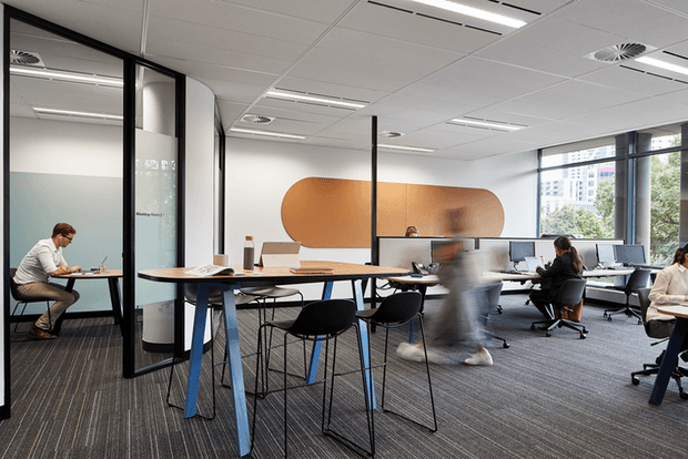 Contemporary open-plan office with shared workstations, individual desks, glass-partitioned meeting rooms, computer monitors, and a large curved copper-toned feature wall. The space has a suspended acoustic ceiling, linear lighting, carpet flooring, and expansive windows overlooking an urban setting.