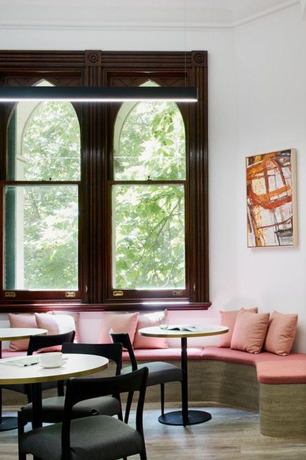 A refined café or commercial lounge interior with high white ceilings, ornate dark timber window frames, large arched windows, pink upholstered banquette seating, round café tables, and black chairs. Light timber flooring and a small abstract artwork add warmth and character.