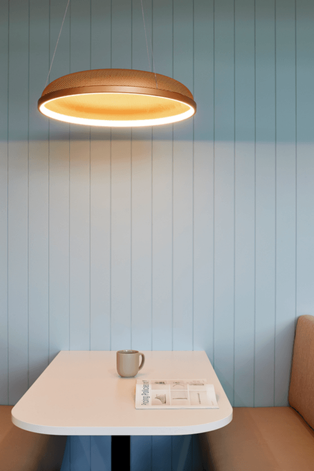A compact contemporary seating nook with a rounded white tabletop, blue pedestal base, upholstered tan bench seating, and a suspended copper-toned oval pendant light. The wall behind is clad in pale blue vertical panelling, creating a calm, softly lit café or lounge setting. A coffee cup and magazine sit on the table.