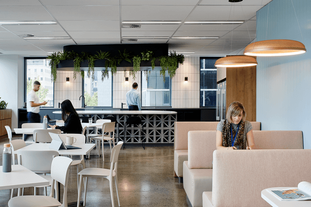 A bright contemporary commercial café or workplace lounge with white tables and chairs, upholstered booth seating, a long counter with geometric breeze-block detailing, hanging pendant lights, and abundant suspended greenery. Large windows overlook an urban setting, while pale blue wall cladding and polished concrete-style flooring create a clean, modern atmosphere.