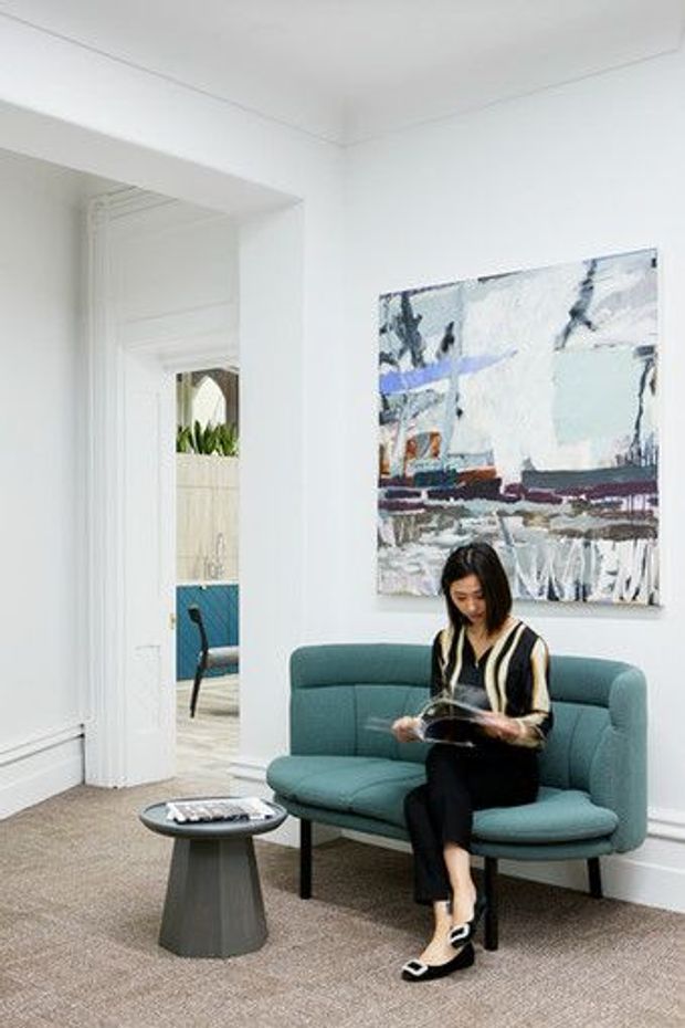 Bright, refined sitting area with white walls, decorative moulding and a large abstract artwork. A teal upholstered sofa and a small dark side table sit on a neutral carpet, with a person reading. An open doorway reveals an adjacent room with a blue seating element and indoor plants.
